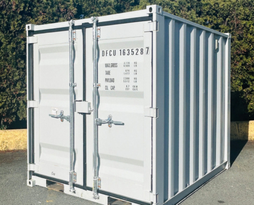 A white shipping container with closed double doors stands on a paved surface outdoors, backed by green trees and a blue sky. The container has identification numbers and weight information printed on the front.