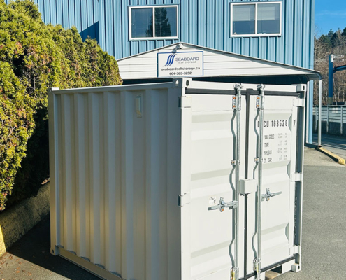 A small, white shipping container is placed on a paved lot in front of a blue industrial building with a Seaboard Self Storage sign and shrubs on the left side.