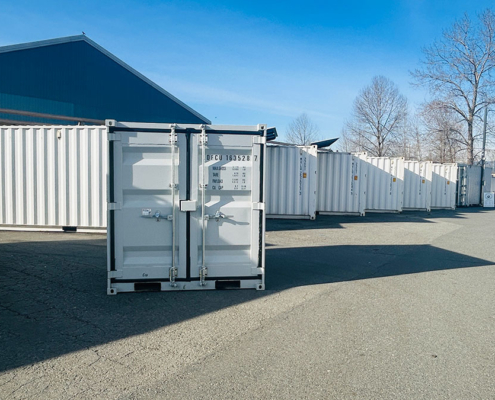 Several white shipping containers are lined up outdoors on a paved lot near a blue building, with bare trees and mountains visible in the background under a clear blue sky.