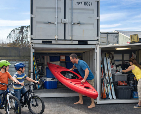 A man takes a red kayak from a storage container as two children with bikes and helmets watch; a woman organizes sports gear inside the container on a sunny day.
