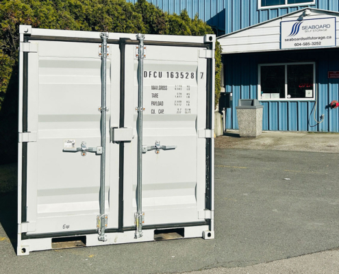 A white shipping container sits on a paved surface outside a blue building with a sign that reads Seaboard Self Storage. Green bushes are visible in the background.