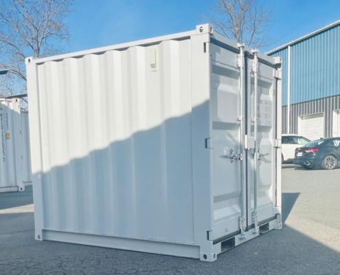 A white shipping container sits on an outdoor lot near a blue industrial building, with a few cars and leafless trees in the background under a clear blue sky.