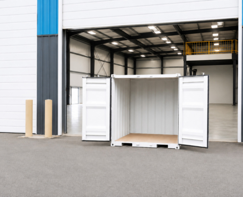 Seasonal-Storage-6-x-7-Container A white shipping container with open doors sits outside a large, open warehouse with blue and gray accents and an empty interior.